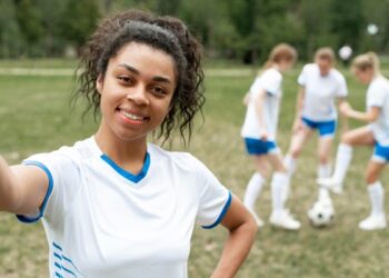 Footballeur souriant prenant un selfie, ses coéquipiers s'entraînant derrière lui.