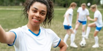 Footballeur souriant prenant un selfie, ses coéquipiers s'entraînant derrière lui.