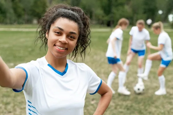 Footballeur souriant prenant un selfie, ses coéquipiers s'entraînant derrière lui.